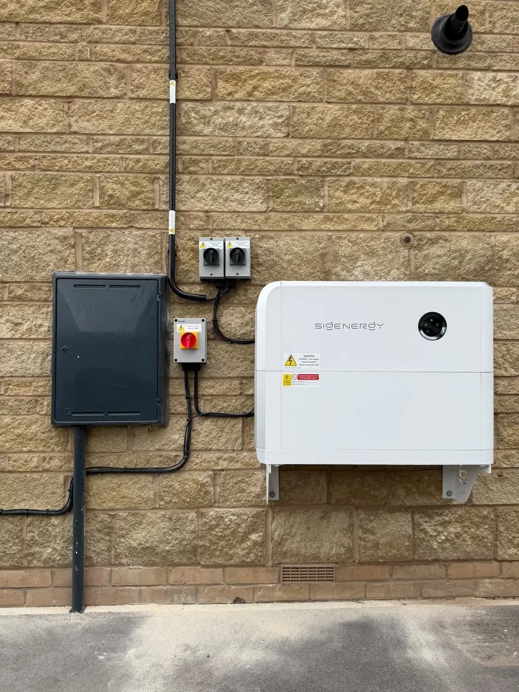 A wall-mounted SigEnergy battery and inverter unit installed on the outside wall of a house, beside its isolator switches and meter box